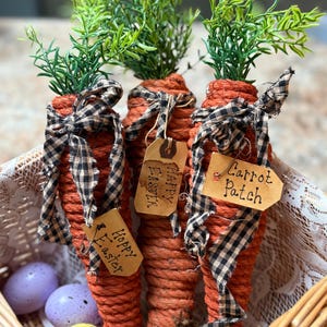 May include: Three orange carrot decorations with green leafy tops, black and white checkered bows, and paper tags that read "Happy Easter" and "Carrot Patch". The carrots are in a woven basket with colorful Easter eggs.