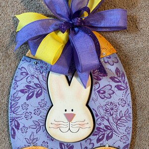 May include: Easter egg-shaped wooden decoration featuring a white bunny face and a purple and yellow bow. The egg has a light purple background with a floral pattern and a gold and orange bottom section with a glittery design. A festive Easter decoration.