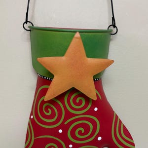 May include: A red metal mitten-shaped Christmas decoration with green swirls and a gold star. The top is green, and a black wire handle is attached for hanging. White dots accent the design.