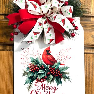 May include: A white, mason jar-shaped Christmas decoration with a red cardinal bird illustration, holly, and the words "Merry Christmas." It features a large red and white bow with cardinal bird accents and red berries.