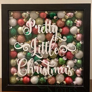 Puede incluir: Una caja de sombra negra llena de una variedad de pequeños adornos navideños coloridos. Los adornos son en tonos de rojo, verde, rosa, blanco y dorado. Las palabras "Pretty Little Christmas" están escritas en letras de vinilo blanco en la parte delantera de la caja.