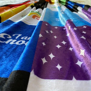 May include: A multi-colored fleece blanket with different patterns and phrases. The blanket features a purple section with white stars, a blue section with the phrase "I am Enough", and a yellow section with a rainbow.