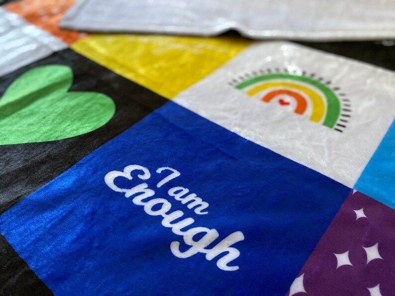 May include: A colorful patchwork quilt with different squares featuring a green heart, a rainbow, and the words "I am Enough" in white on a blue square.