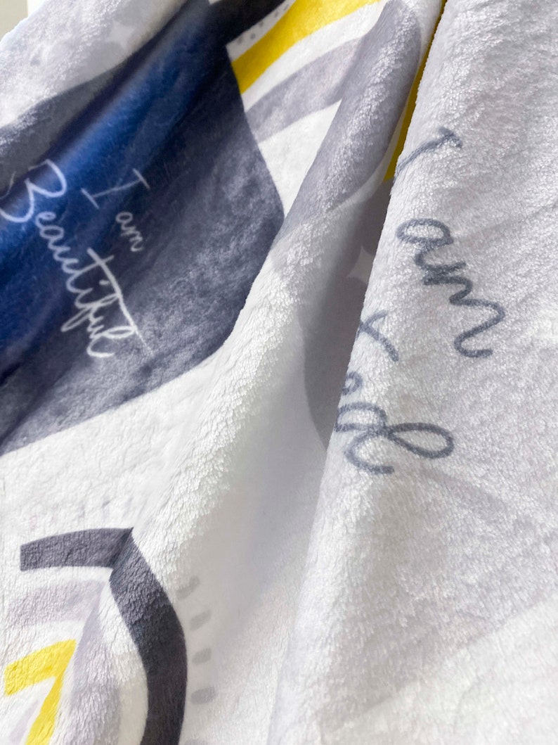 May include: A white, gray, and yellow fleece blanket with the text "I am Beautiful" printed on it.