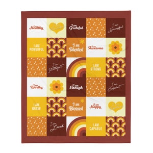 May include: A colorful throw blanket with a patchwork design featuring positive affirmations like "I am strong", "I am worthy", and "I am loved". The blanket is made up of squares with different patterns and colors, including yellow, orange, brown, and white.