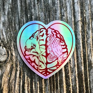 May include: A heart-shaped sticker with a holographic finish. The sticker features a red outline of a woman pole dancing on the left side and a pair of hands in prayer on the right side.