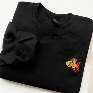 Könnte beinhalten: Schwarzer Pullover mit einem gestickten orangefarbenen und gelben Fischdesign.