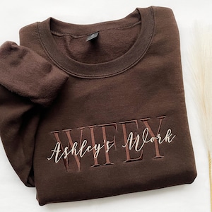May include: A dark brown crewneck sweatshirt featuring the embroidered text WIFEY in a bold serif font with the name Ashley's Work layered over it in a white cursive script. The garment has a ribbed collar and long sleeves with folded cuffs.