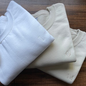 Puede incluir: Tres sudaderas plegadas en diferentes tonos de beige, azul claro y blanco. Cada sudadera tiene una sola letra bordada en la parte delantera: L, O y M.