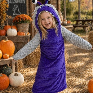 May include: A child in a purple monster costume with a hood and large eyes, smiling. The costume has gray sleeves and a long, draped body. The background includes pumpkins, a sign that says "Happy Halloween", and fall foliage.