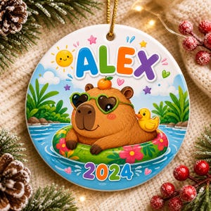 May include: A round ceramic ornament featuring a cartoon capybara in a pool float with heart-shaped sunglasses. The ornament has the name "ALEX" and the year "2024" printed on it. A yellow duck and a smiling sun are also featured.