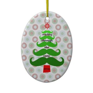 May include: Oval Christmas ornament with a green mustache tree design and the words "staching through the snow". The ornament has a white background with red and green polka dots and a yellow hanging string. The year 2013 is printed on the bottom.