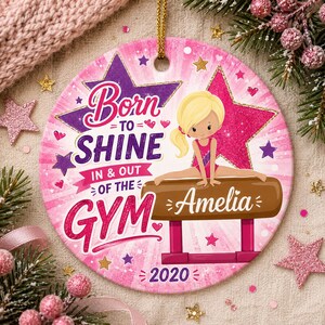 May include: A pink and white ceramic ornament with a gymnast illustration. The ornament has the text "Born to Shine In & Out of the Gym" and the name "Amelia". The year "2020" is also displayed.