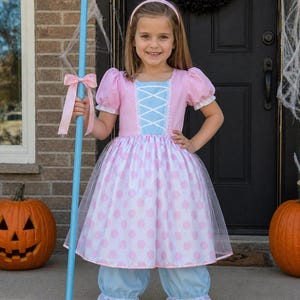May include: A child's costume featuring a pink and white polka-dot dress with a blue bodice and bloomers. The costume includes a light blue shepherd's crook with a pink bow. Two carved pumpkins are on the porch.