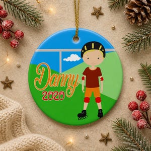 May include: Round ceramic ornament featuring a cartoon illustration of a boy in roller skates. The ornament has a blue sky background with a green field. The name "Danny" and the year "2020" are written in orange and green. A gold string is attached for hanging.