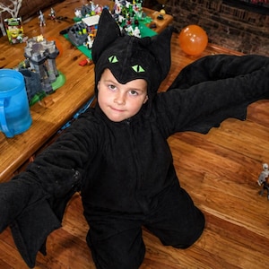 May include: A child in a black bat costume with a hood and wings, posing on a wooden floor. The costume has green eye details on the hood. Halloween decorations, including toy figures and a spider, are scattered around.