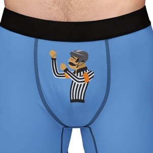 May include: Pale blue boxer shorts with a black waistband. The shorts have a cartoon referee graphic in black, white, and orange. The referee is wearing a black hat and a striped shirt.