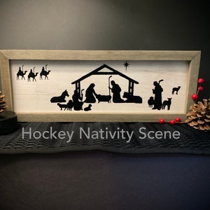 May include: A wooden framed sign with a white background and a black silhouette of the nativity scene. The sign reads "Hockey Nativity Scene".