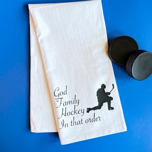 May include: White kitchen towel with black text that reads "God Family Hockey In that order" and a black silhouette of a hockey player.