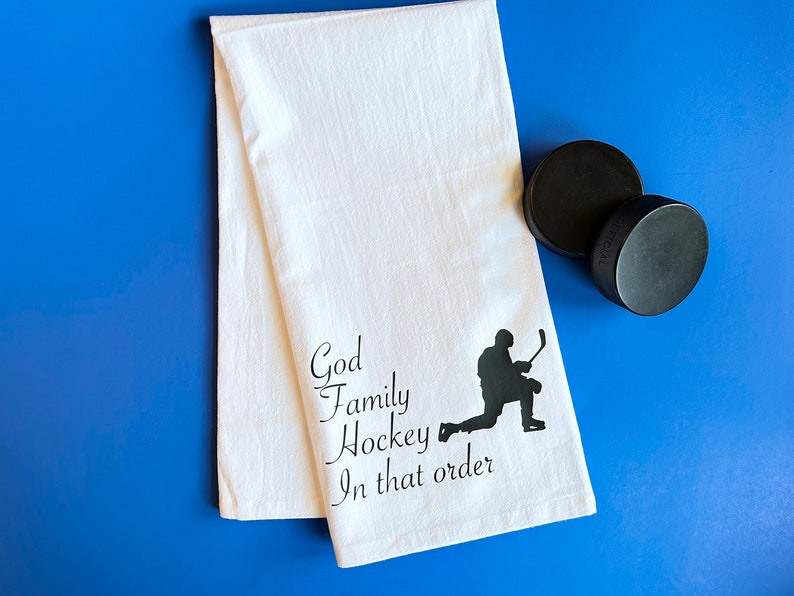 May include: White kitchen towel with black text that reads "God Family Hockey In that order" and a black silhouette of a hockey player.