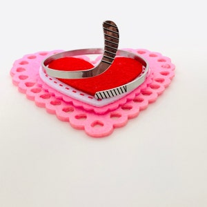 May include: A silver hockey stick charm bracelet on a pink and white heart-shaped felt background.