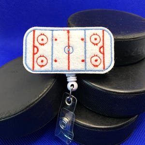May include: A white and blue hockey rink badge reel with a clear plastic clip. The badge reel features a hockey rink design with red and blue lines and circles. The background includes black hockey pucks.