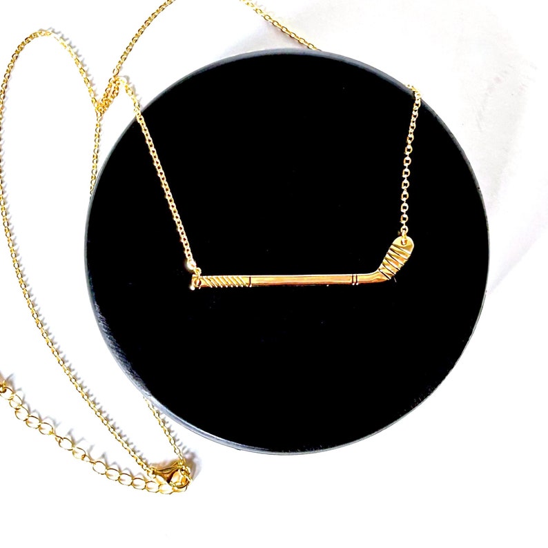 May include: A gold necklace with a hockey stick pendant. The necklace is on a black velvet background.