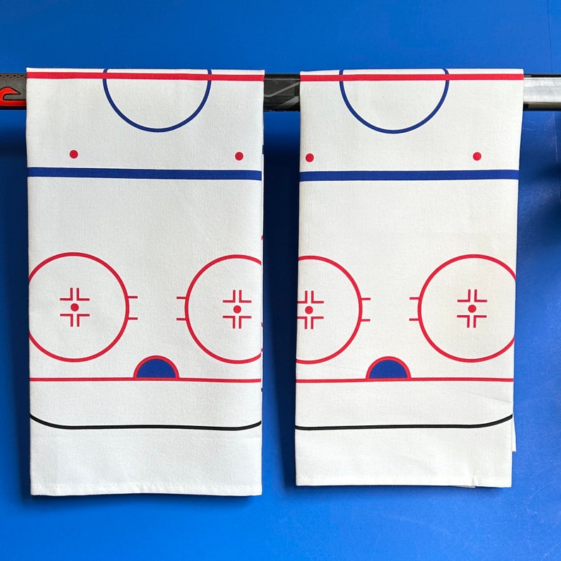 May include: Two white kitchen towels with a hockey rink design printed on them. The rink is outlined in red and blue, with a center circle and two face-off circles. The towels are hanging on a metal bar.