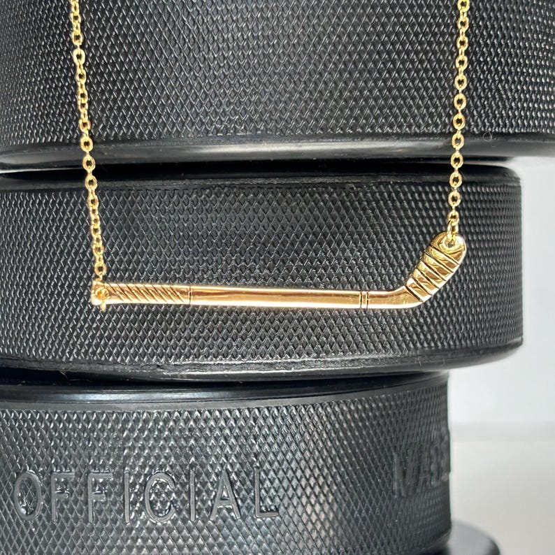 May include: A gold hockey stick necklace on a black background. The necklace is made of a thin gold chain and a small hockey stick pendant. The hockey stick is detailed with a curved blade and a straight shaft.