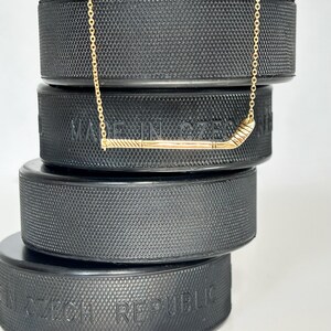 May include: A gold necklace with a hockey stick pendant. The necklace is displayed on top of hockey pucks.