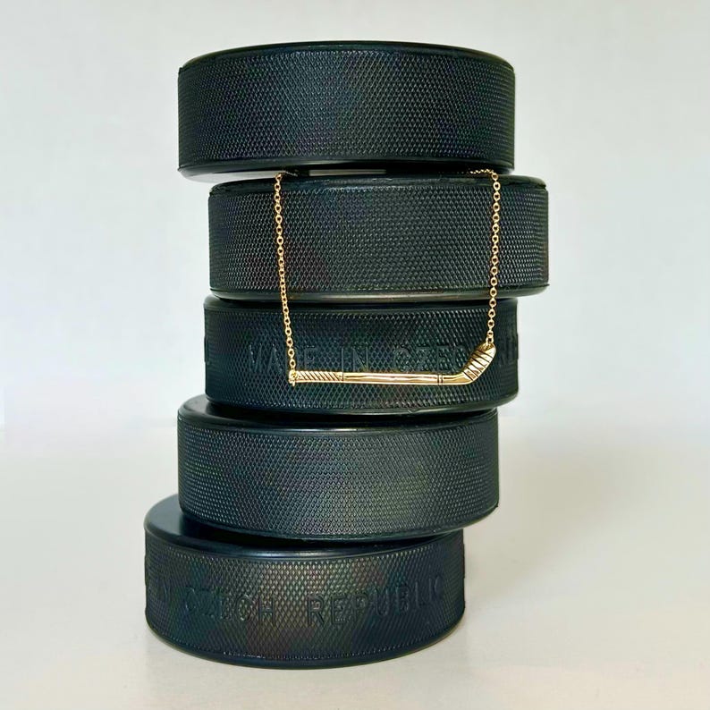 May include: A gold hockey stick necklace on a stack of black hockey pucks. The pucks are stacked on top of each other and the necklace is draped over the top puck.