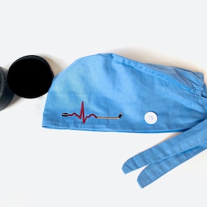 May include: A light blue surgical scrub hat with a red and black embroidered heartbeat and hockey stick design. The hat has a white button on the side and two long ties.