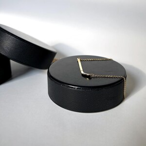 May include: A gold hockey stick necklace with a small diamond on the blade, displayed on a black hockey puck.