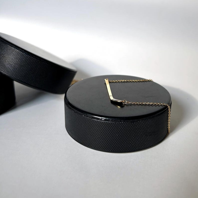 May include: A gold hockey stick necklace with a small diamond on the blade, displayed on a black hockey puck.