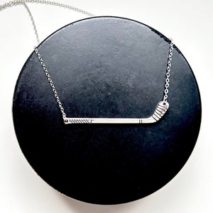 May include: A silver necklace with a hockey stick pendant. The pendant is a simple design with a slight curve at the end. The necklace is on a black surface.