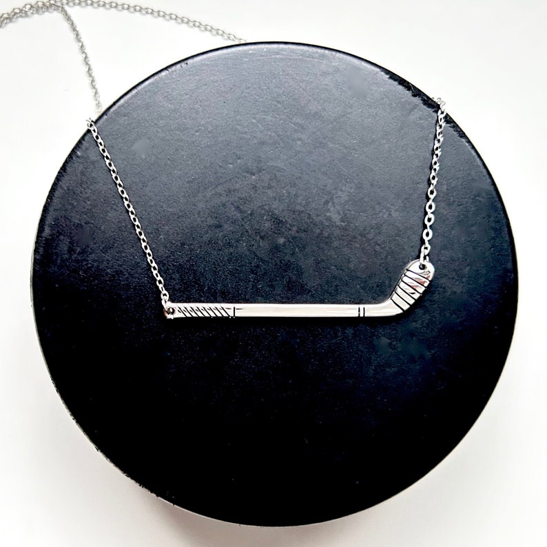 May include: A silver necklace with a hockey stick pendant. The pendant is a simple design with a slight curve at the end. The necklace is on a black surface.