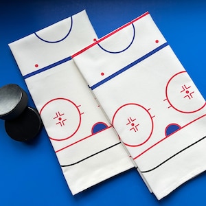 May include: Two white cotton tea towels with a hockey rink design. The rink is outlined in black and features red and blue details. The towels are folded and displayed on a blue surface.