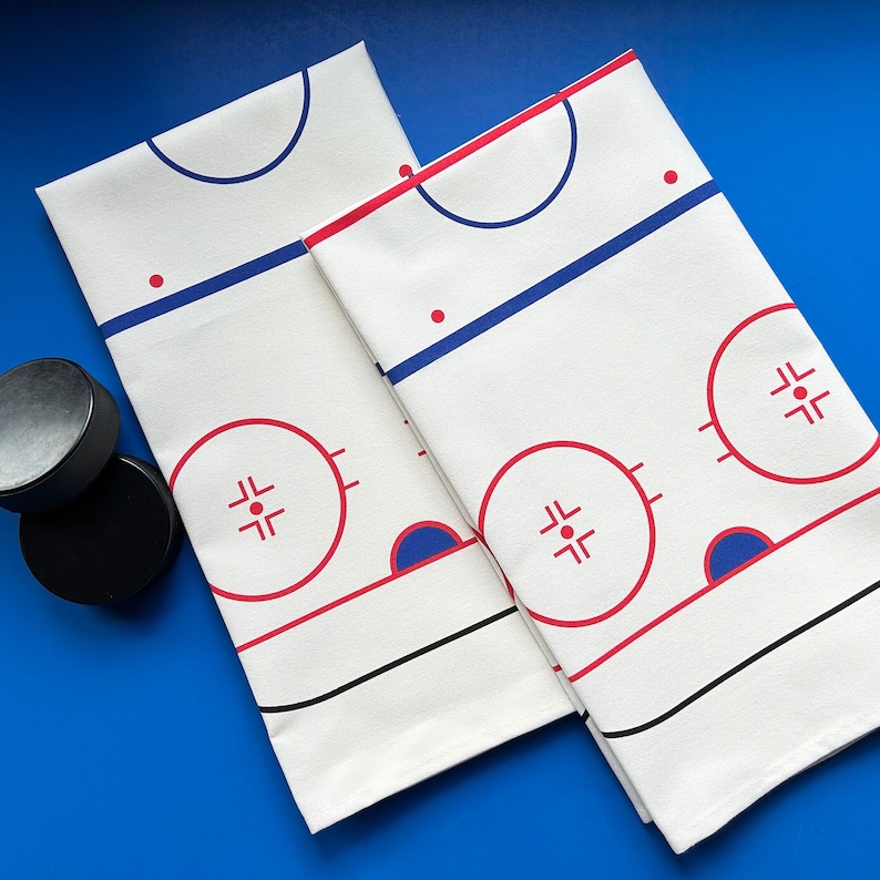 May include: Two white cotton tea towels with a hockey rink design. The rink is outlined in black and features red and blue details. The towels are folded and displayed on a blue surface.