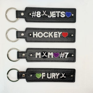 May include: Four black leather keychains with silver key rings. Each keychain has a different design with rhinestones and colored hearts. The first keychain has a hockey stick and the text '#8 JETS'. The second keychain has the text 'HOCKEY'. The third keychain has a hockey stick and the text 'MOM #7'. The fourth keychain has a hockey stick and the text 'FURY'.
