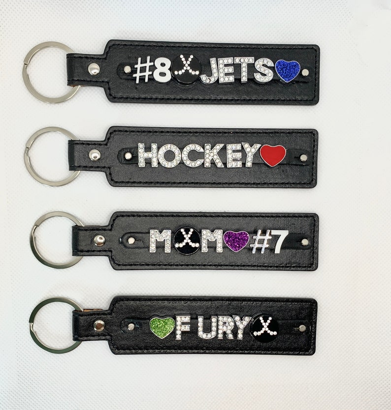 May include: Four black leather keychains with silver key rings. Each keychain has a different design with rhinestones and colored hearts. The first keychain has a hockey stick and the text '#8 JETS'. The second keychain has the text 'HOCKEY'. The third keychain has a hockey stick and the text 'MOM #7'. The fourth keychain has a hockey stick and the text 'FURY'.