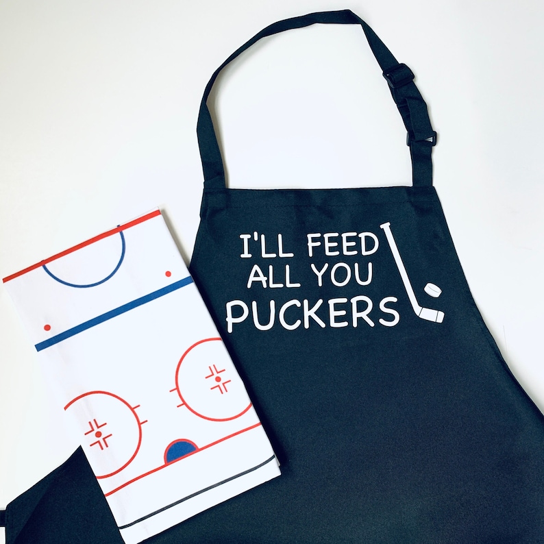 May include: Black and white kitchen apron with the text "I'll feed you all you puckers" and a hockey stick and puck graphic. A white and blue hockey rink design is on a white tea towel.