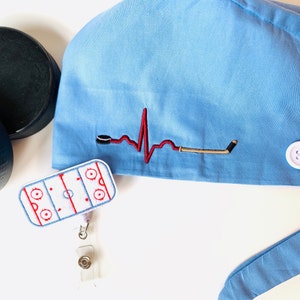 May include: Light blue scrub cap with a hockey-themed embroidered design. Features a red heartbeat line with a hockey puck and stick. Includes a hockey rink badge reel. Two black hockey pucks are visible.