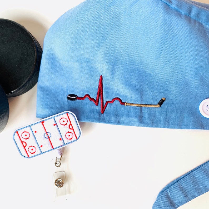 May include: Light blue scrub cap with a hockey-themed embroidered design. Features a red heartbeat line with a hockey puck and stick. Includes a hockey rink badge reel. Two black hockey pucks are visible.