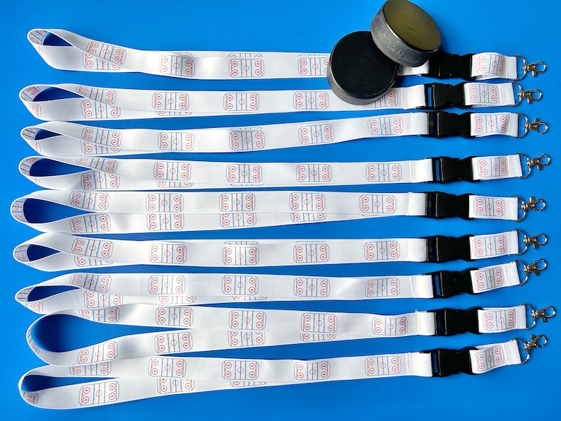 May include: Ten white lanyards with a hockey rink design printed on them. Each lanyard has a black breakaway clip and a silver swivel hook. Two black hockey pucks are also included in the image.