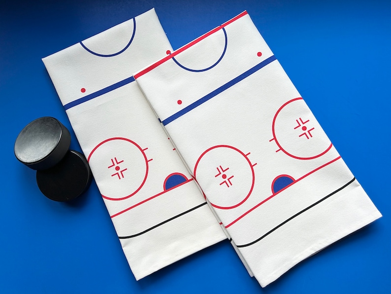 May include: Two white cotton dish towels with a hockey rink design. The rink is outlined in black and red, with blue and red accents. The towels are folded and placed on a blue surface.