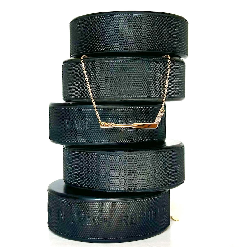May include: A gold hockey stick necklace on a stack of black hockey pucks. The pucks have the words "Made in Czech Republic" printed on them.