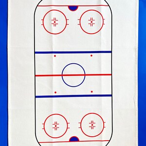 May include: White cotton kitchen towel with a black and red outline of a hockey rink. The rink has two blue lines, two red lines, two face-off circles, and a center circle.
