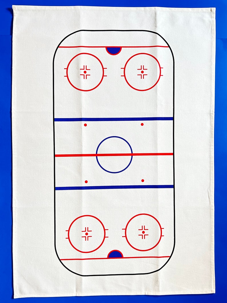 May include: White cotton kitchen towel with a black and red outline of a hockey rink. The rink has two blue lines, two red lines, two face-off circles, and a center circle.