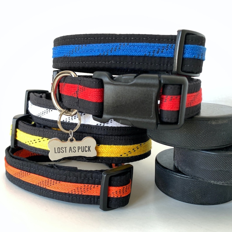 May include: Four dog collars stacked on top of each other. The collars are black with different colored stripes. The top collar has a blue stripe, the second collar has a red stripe, the third collar has a white stripe, and the bottom collar has a yellow stripe. A silver dog tag with the text "LOST AS PUCK" is attached to the third collar.