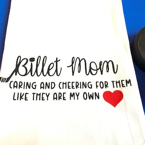 May include: White kitchen towel with black text that reads "Billet Mom Caring and Cheering for Them Like They Are My Own" with a black hockey stick and a red heart.